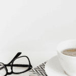A pair of black glasses and a cup of coffee on a white surface.