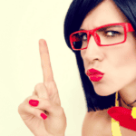 Woman with black hair and red glasses making a 'shush' gesture.