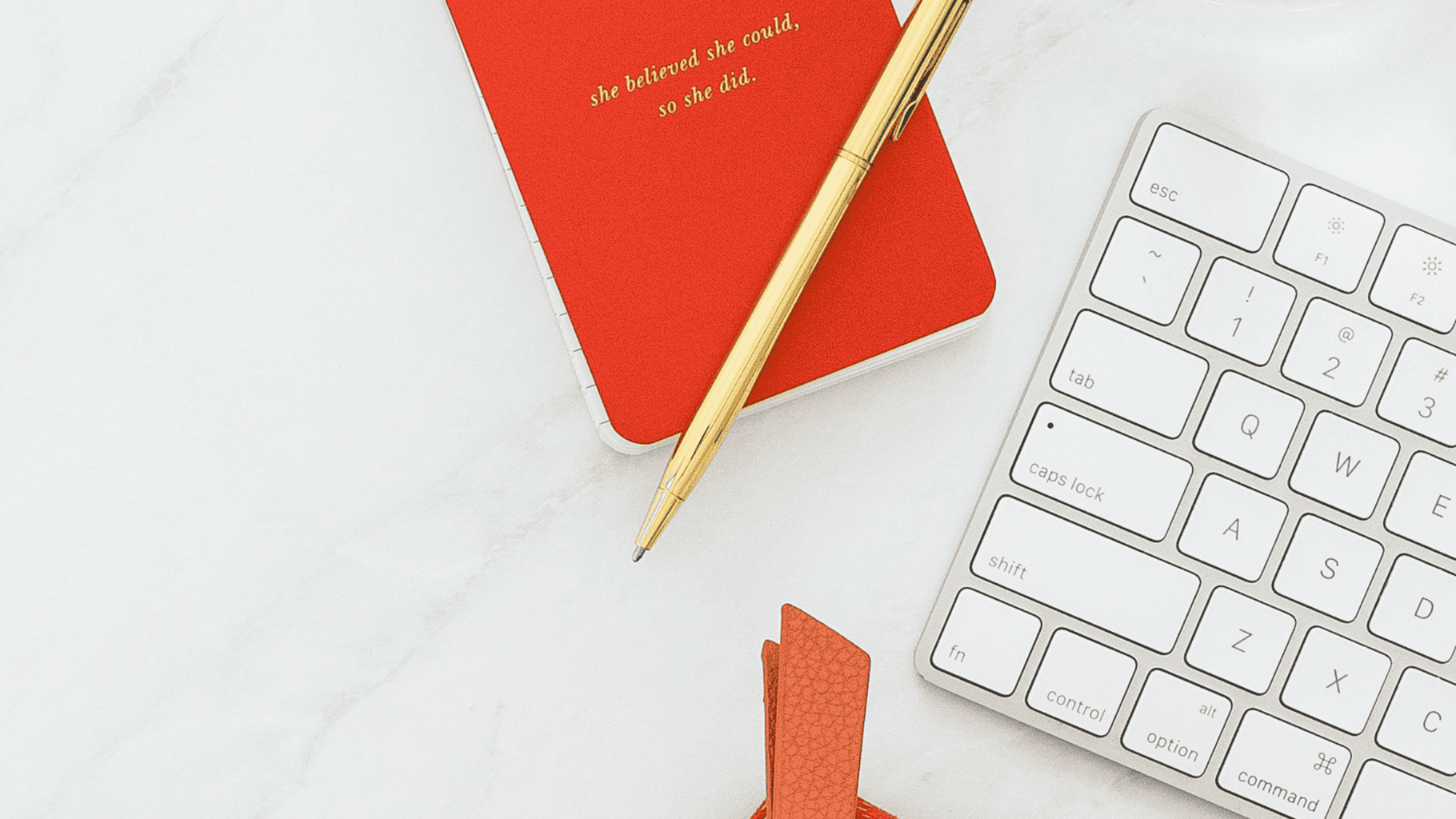 Red notebook with gold pen and white keyboard on marble surface.