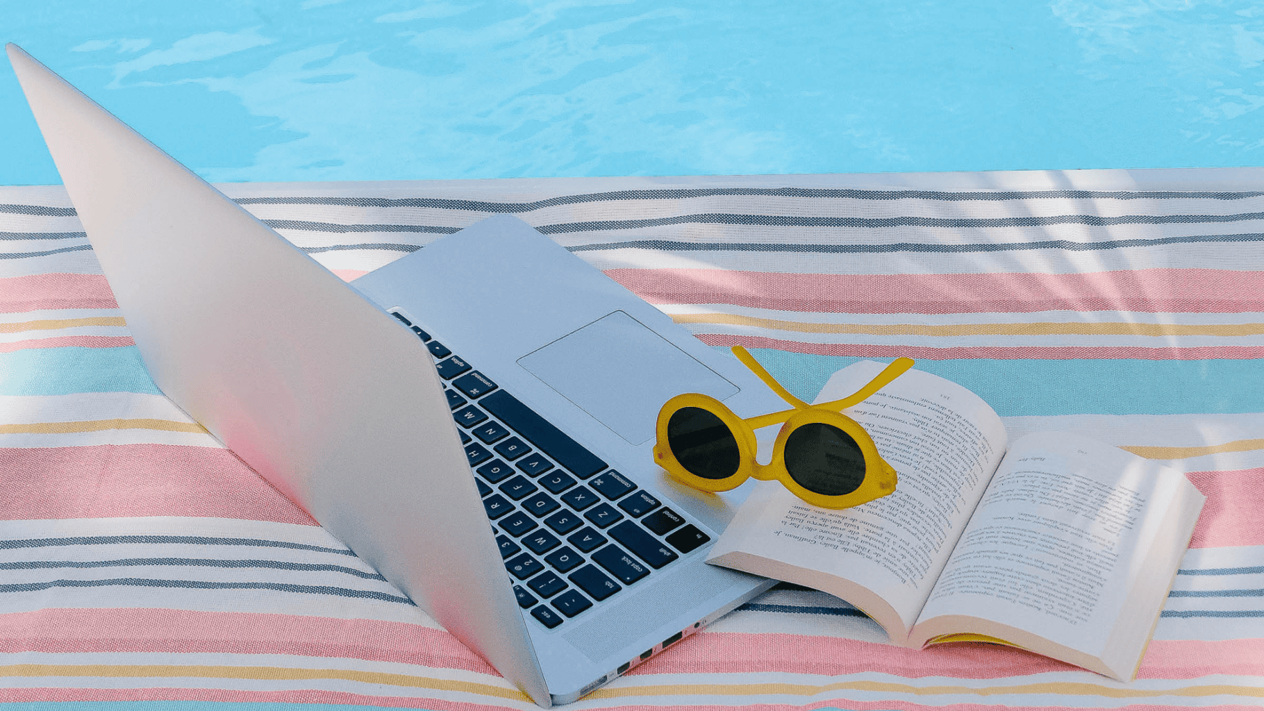 Laptop, sunglasses, and open book on a striped towel by the pool.