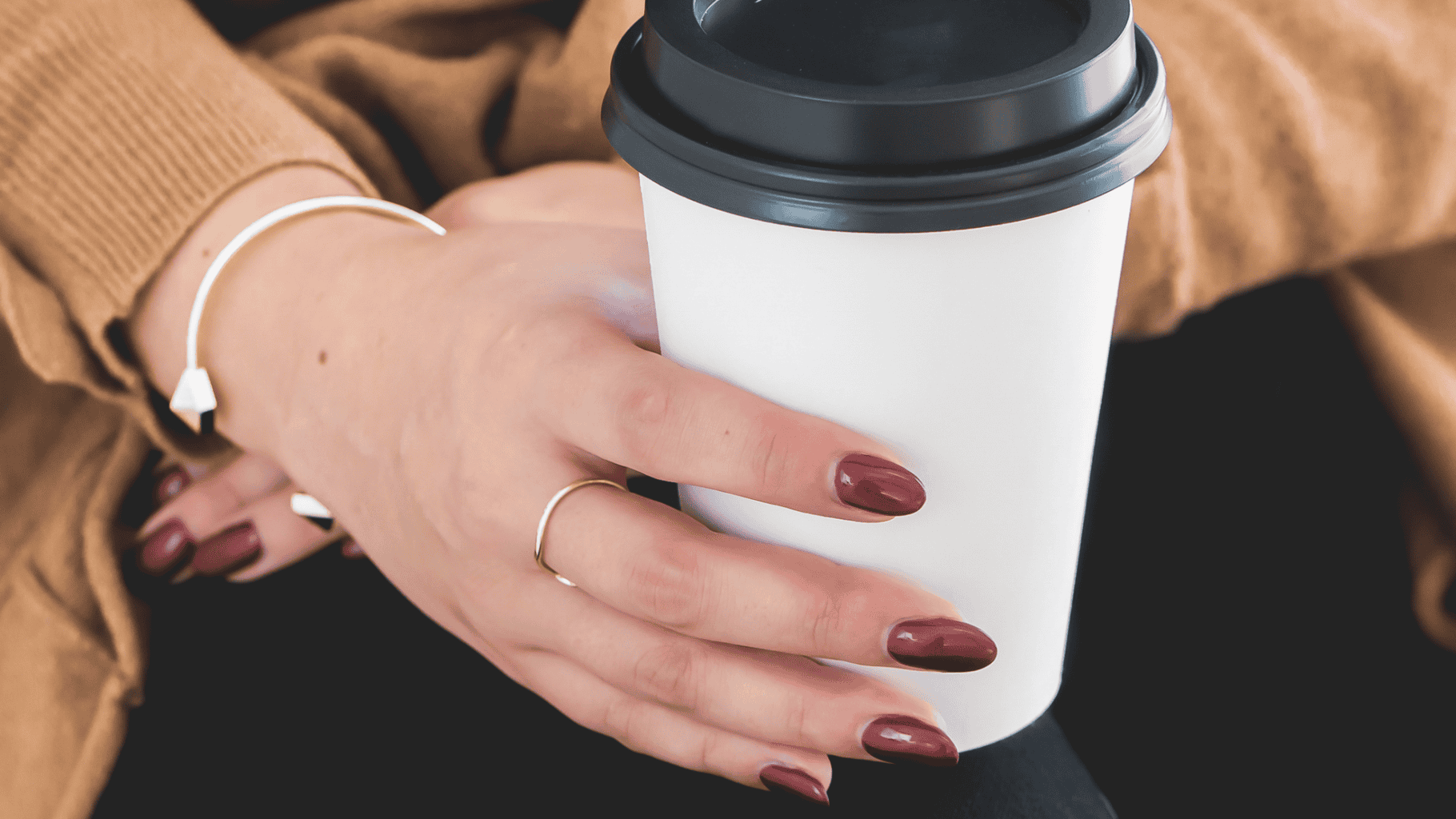 Close-up of a woman holding a coffee cup with manicured nails and a brown sweater.